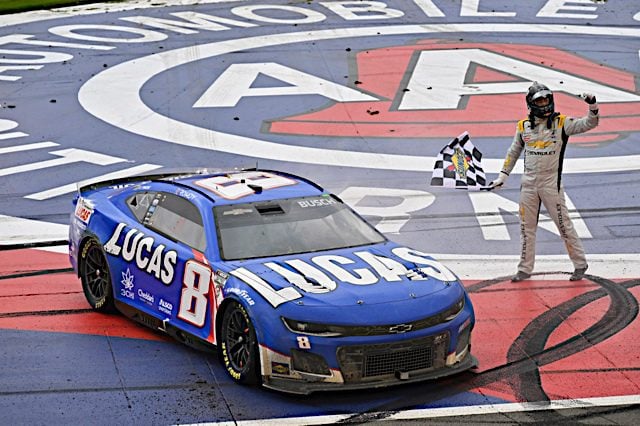 Kyle Busch holding the checkered flag next to his car after winning the NASCAR Cup Series race at Auto Club Speedway, NKP